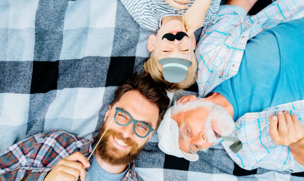 Grandfather,father and son enjoying together lying on a checkered blanket. Tree men of different ages smiling playing with fake mustache, hat, glasses. Top view of boy and his dad, granddad with smile
