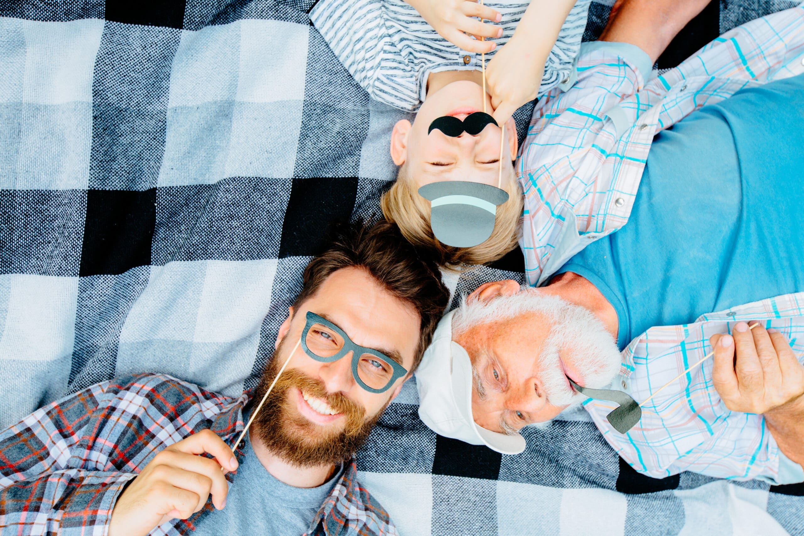 Grandfather,father and son enjoying together lying on a checkered blanket. Tree men of different ages smiling playing with fake mustache, hat, glasses. Top view of boy and his dad, granddad with smile